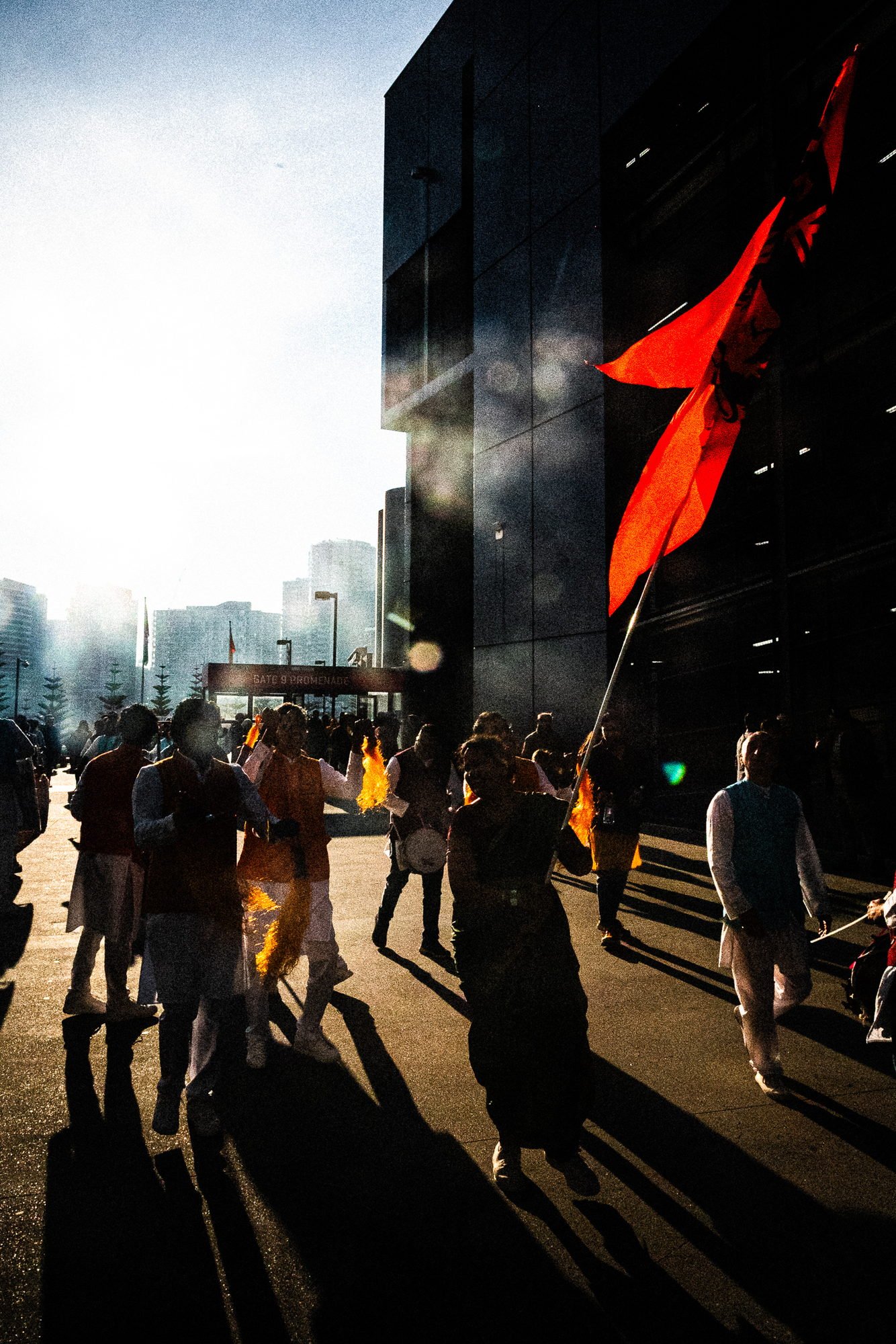 Saffron flag against the black glass of the stadium, low sun and haze, figures walking in silhouette