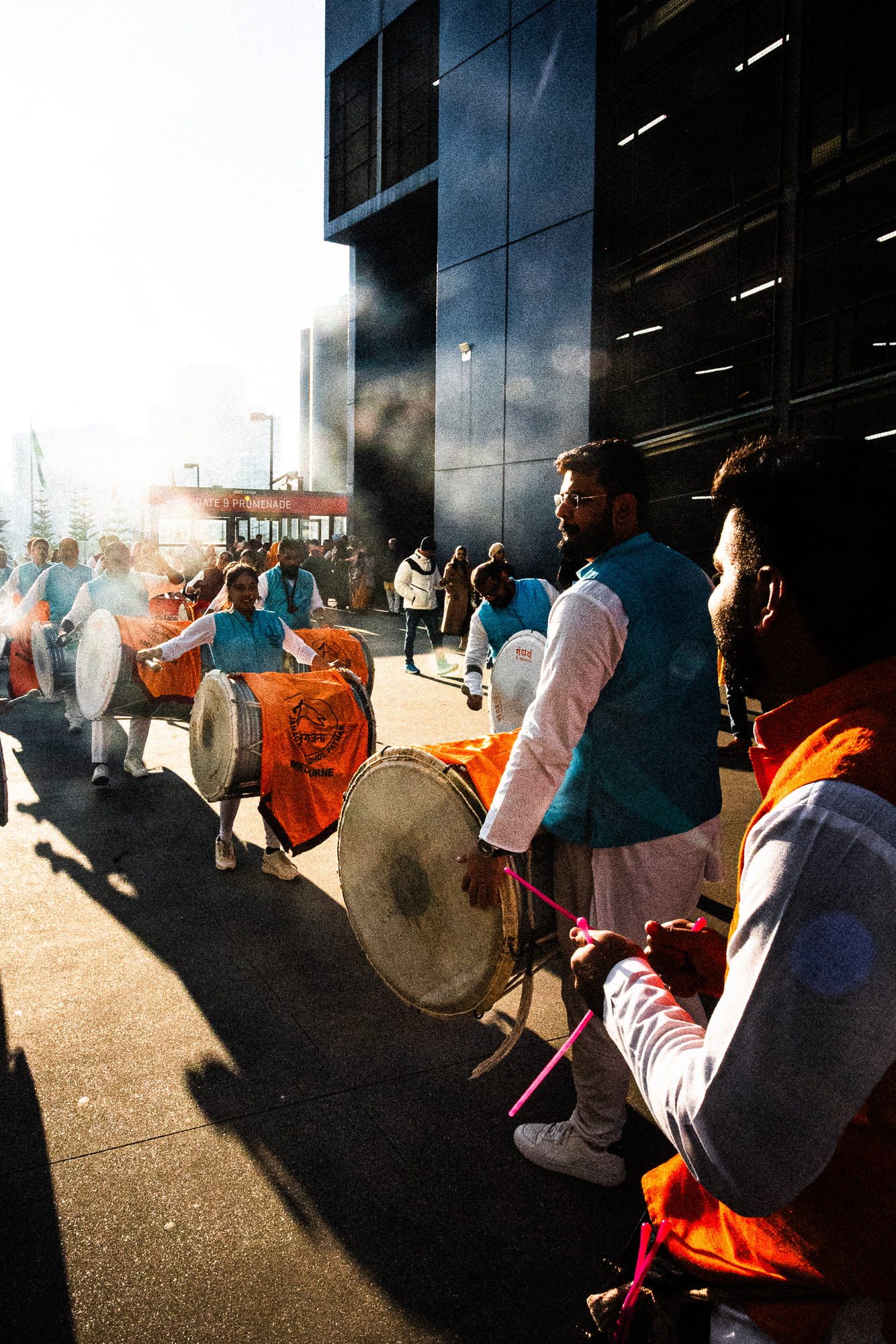Dhol players in blue vests walking away through drum smoke, long shadows across the concourse