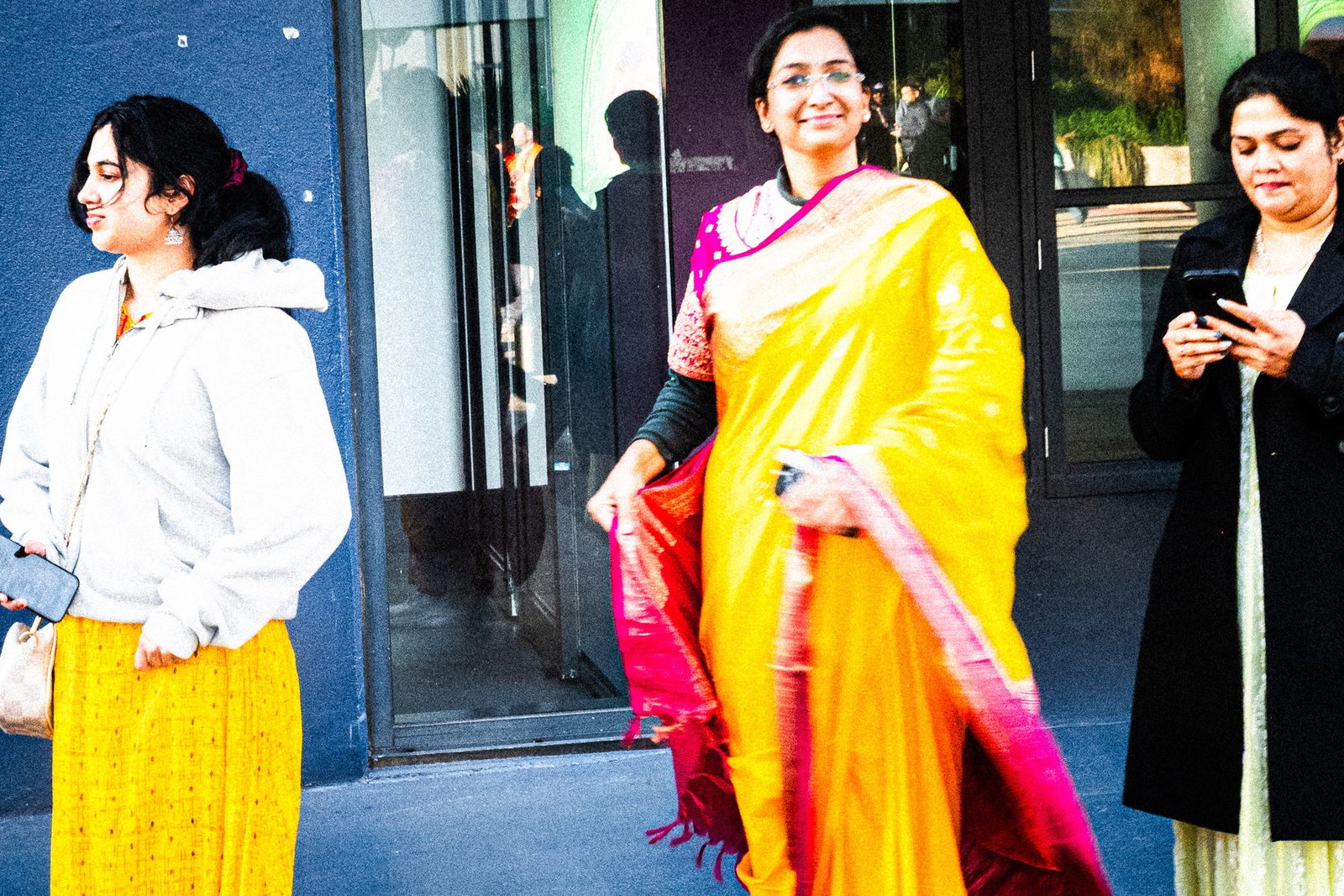 Three women against a blue and purple wall, one on her phone, one in a yellow sari