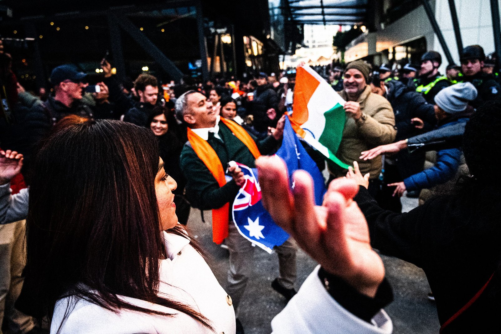 A blurred hand sweeps across the frame, flags and a woman's profile behind it, police at the far edge